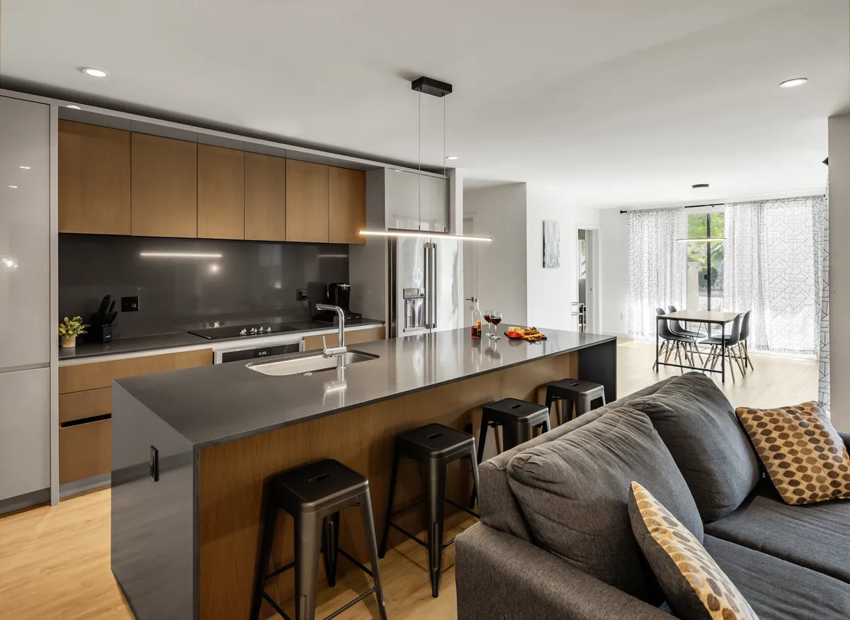 The image showcases a contemporary, open-concept living space featuring a stylish kitchen island with modern stools, a cozy couch, and an inviting dining area. The setting is designed for cooking, entertaining, and relaxing, all bathed in natural light, emphasizing comfort and upscale finishes.