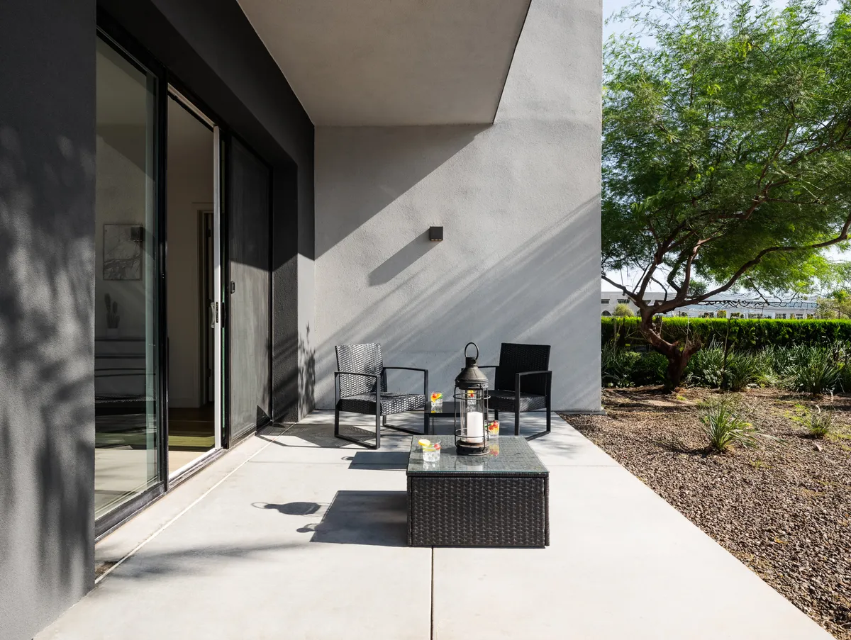 A serene exterior view of a landscaped outdoor seating area with a coffee table, lantern, and lush garden setting. The space is inviting for having coffee, enjoying a book, or socializing with colleagues after work.