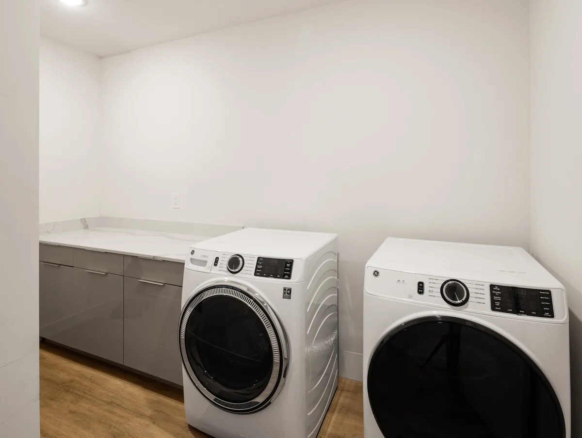 A bright and organized laundry area featuring a front-loading washing machine and dryer set beneath a spacious countertop, with cabinets above for extra storage. The space is clean and practical, reflecting an efficient setup ideal for longer stays or families.