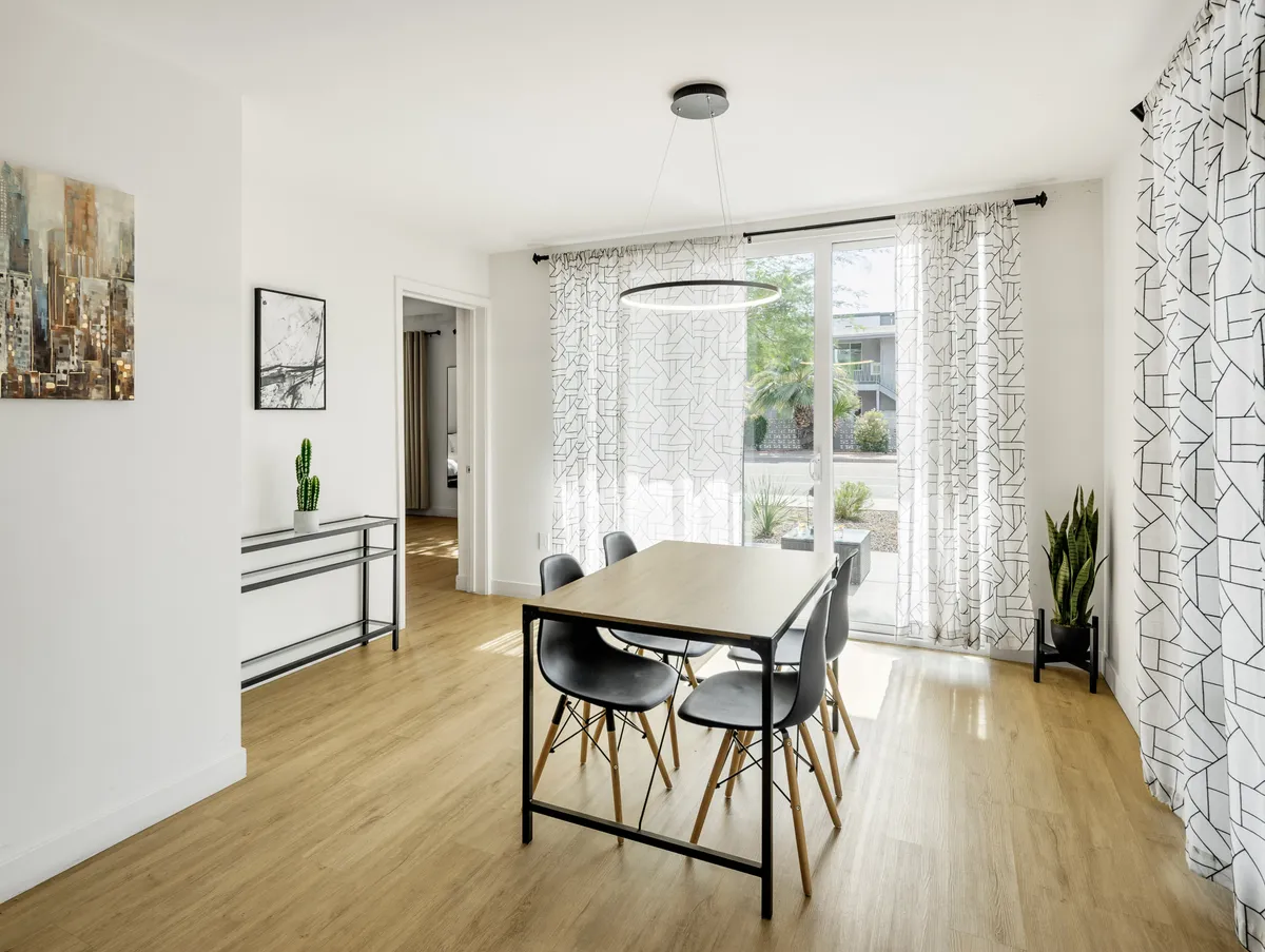 A contemporary dining space with a large table, elegant chandeliers, expansive windows letting in natural light, and stylish decorative art. Potted plants and comfortable seating create a welcoming atmosphere for families or groups.