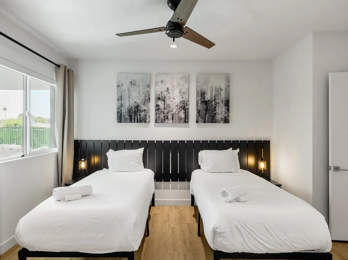 The room showcases two twin beds with a modern black wooden headboard and abstract artwork on the wall. Natural light fills the space, and the window offers a view outside, creating a tranquil, airy feel.