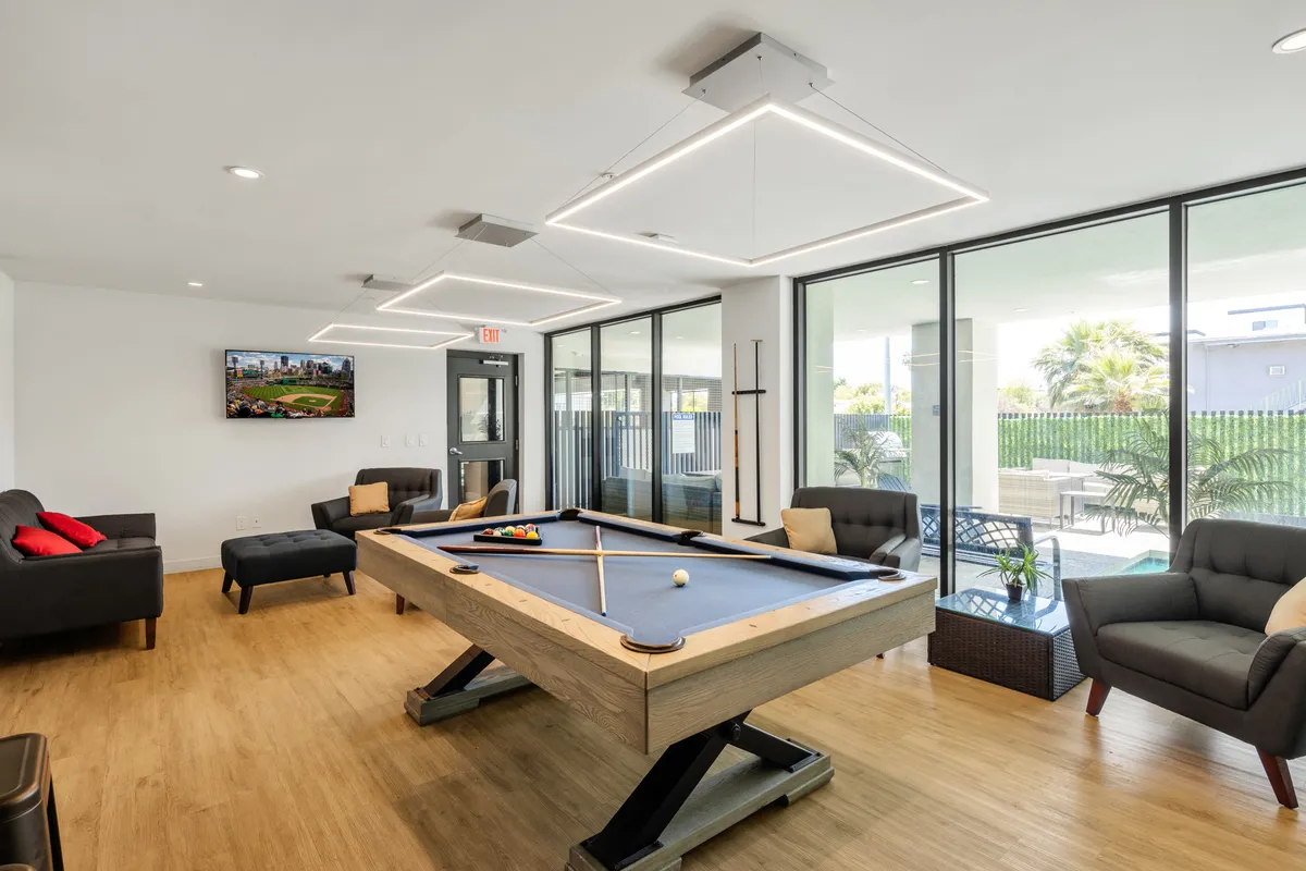 A spacious and inviting clubhouse interior featuring a pool table at its center, surrounded by comfortable seating and large windows that allow natural light to brighten the space. The room also includes a television, creating a lively environment for social gatherings, games, and relaxation.