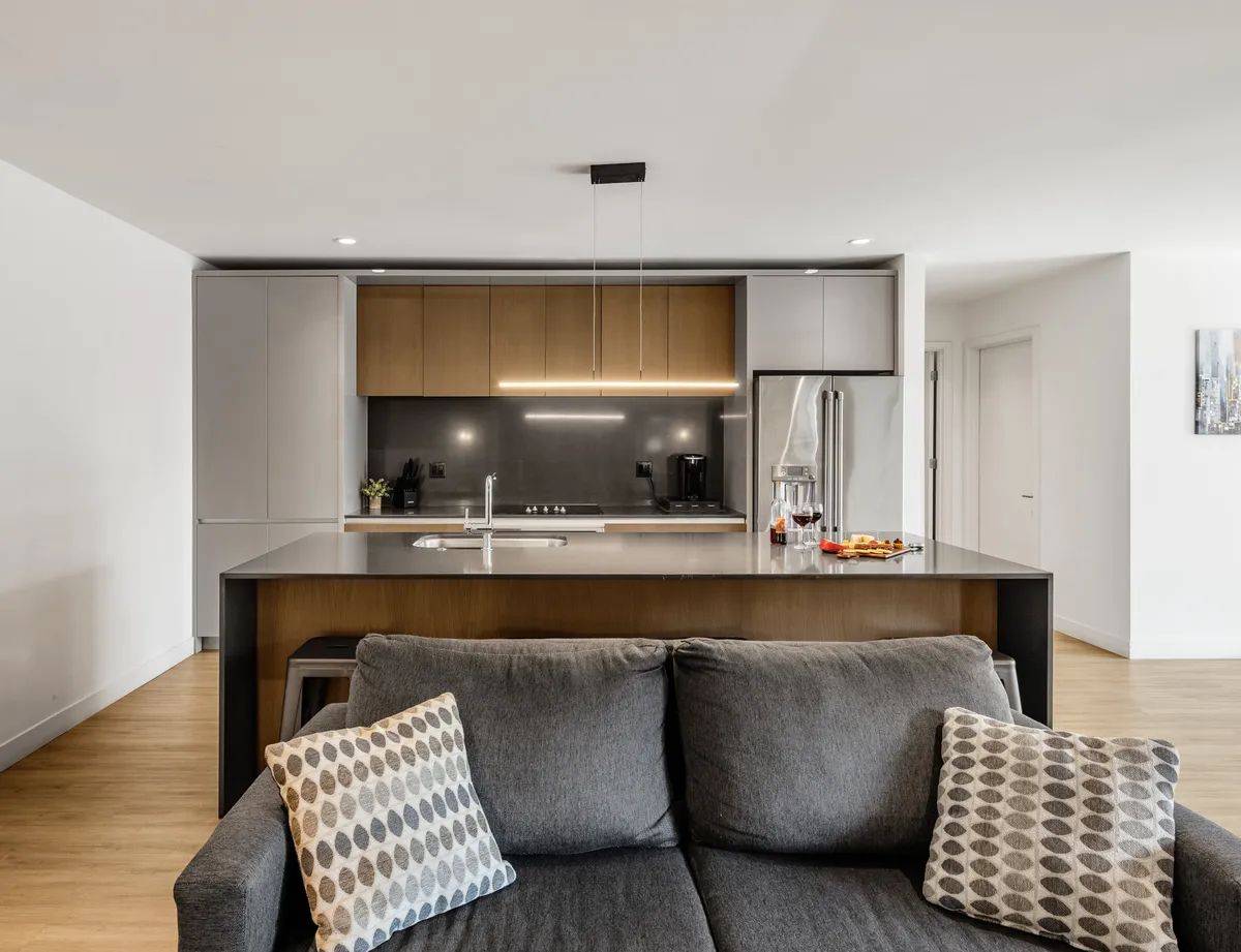 This image offers a sweeping view of the open-concept kitchen and living area. The modern kitchen island, premium appliances, and inviting sofa are bathed in natural light, with stylish decorative accents and cabinetry that create a contemporary, cozy ambiance. The image highlights both the social kitchen and the seamless connection to the living zone.