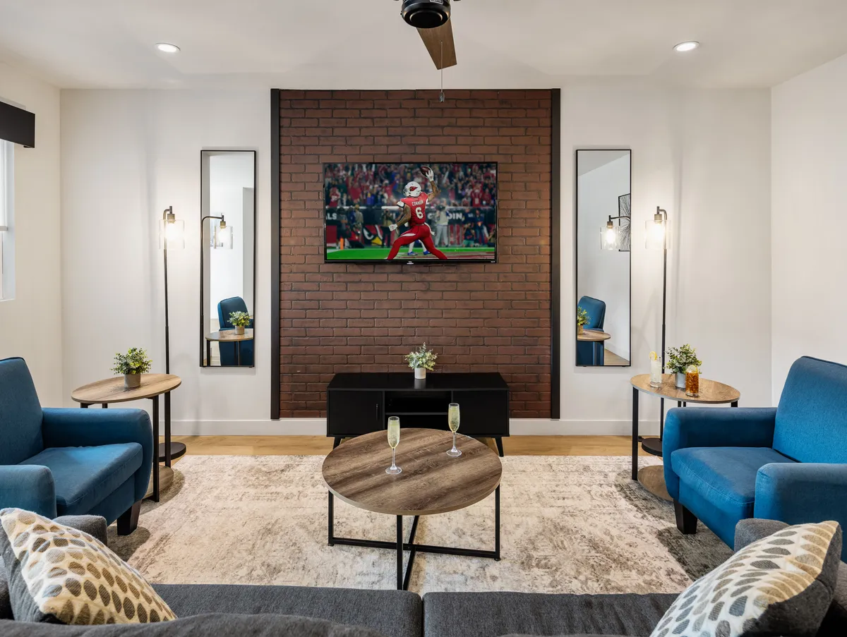 A chic living space featuring a brick accent wall with a smart TV, plush blue sofas, and a coffee table adorned with drinks and games. The room exudes a casual but stylish vibe ideal for game nights, movie marathons, and relaxed socializing.