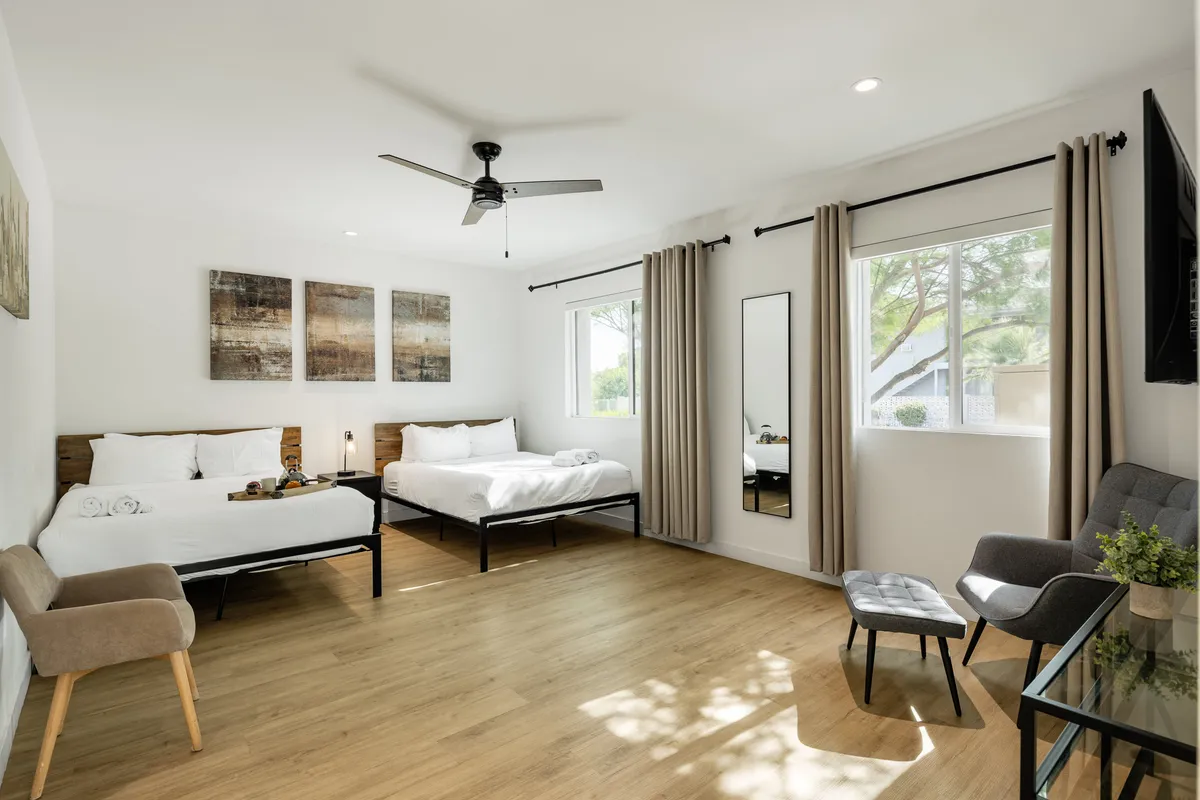 A tranquil bedroom with two beds dressed in crisp white linens, accented by artwork, a mirror, a chair, and a small table by large windows. The space is inviting and adaptable for different guest groupings, with ample natural light.