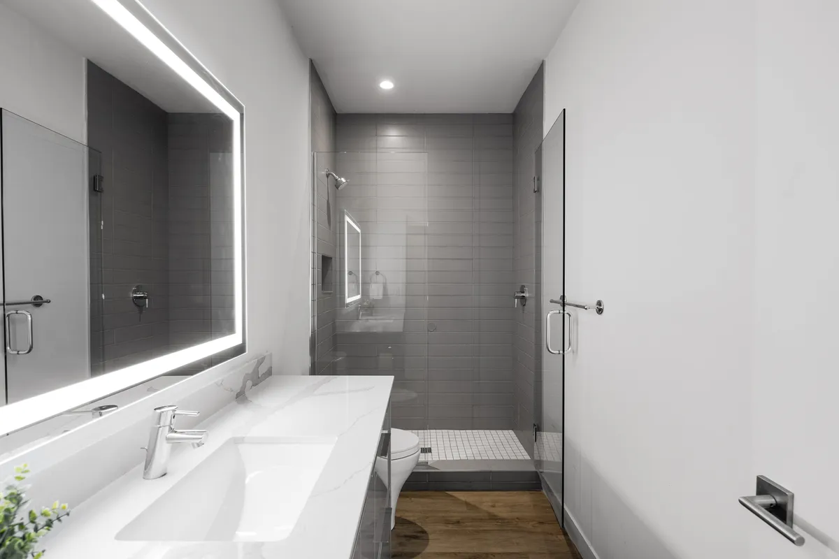 A modern en-suite bathroom featuring a large backlit mirror, sleek sink and faucet, and a glass shower enclosure. The minimalist design and morning light create a spa-like, calming environment.